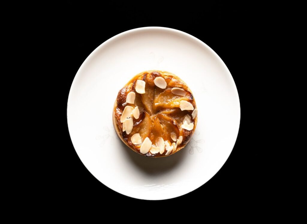 A delectable almond tart with sliced almonds sits elegantly on a white plate against a black backdrop.