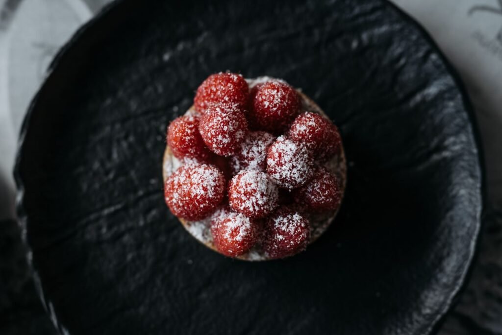 A vibrant raspberry tart with powdered sugar on a textured black plate.